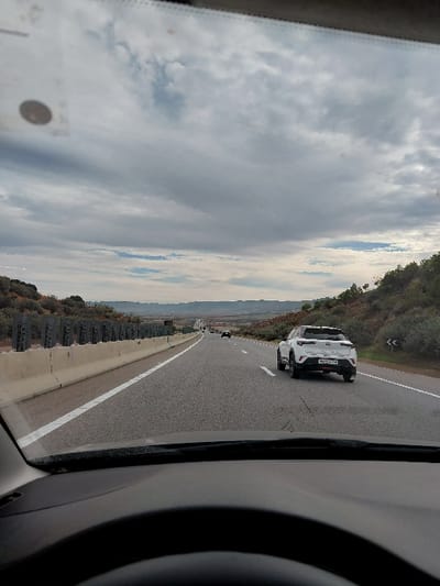 Captured from inside a car, the scene shows a highway stretching into the distance under a cloudy sky. A white SUV is visible in the adjacent lane, and other vehicles are further ahead. A guardrail lines the left side of the road. The car's dashboard and frame partially obstruct the view.