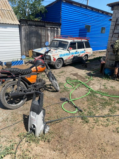 In Mamontovo, Russia, a sunlit outdoor scene unfolds with a yard containing a white station wagon adorned with colorful decals and text in Cyrillic, a motorcycle with an orange seat, and a silver and black pressure washer. The station wagon, possibly a Lada, has a roof rack and a phone number displayed on its rear quarter panel. The motorcycle, a GS model, is parked in front of the car, and a black helmet with red trim rests on its handlebars. A green garden hose snakes across the dirt and patchy grass, connecting to the pressure washer. A person in camouflage clothing stands near a brick structure with a ladder on the right side of the image, facing away from the camera. Behind the car and motorcycle, a weathered wooden fence separates the yard from structures with corrugated metal siding, one of which is bright blue. The ground is a mix of dry dirt, gravel, and sparse vegetation.