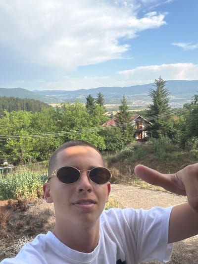 This is a selfie of a young person wearing a white t-shirt and round, gold-rimmed sunglasses. The subject is looking at the camera, with their face partially obscured by a hand raised in a gesture. The background shows a scenic view of rolling hills covered in lush green trees, with a small building nestled among them. In the distance, a valley with fields and some structures can be seen. The sky is blue with scattered white clouds. The overall impression is of a casual outdoor setting, likely during daylight hours with pleasant weather, possibly in a rural or mountainous area. The location context suggests this might be Vladislavtsi, Bulgaria. There is no visible text in the image.
