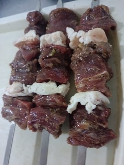 Three skewers of uncooked meat and fat alternate along their length. The meat is a dark red and appears to be marinated. The fat is white and interspersed between the meat pieces. The skewers are made of metal and appear to be resting on a white surface. The skewers are arranged side by side.
