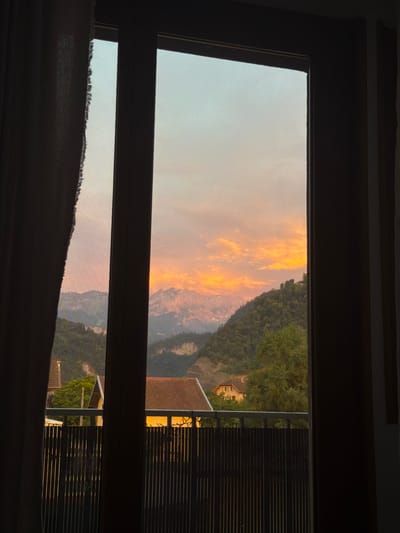 This media file shows a view through a window during sunset. The sky is filled with dramatic clouds in shades of orange and yellow, indicating a vibrant sunset. The scene outside the window features a mountainous landscape covered in dense green trees. In the distance, a few buildings with reddish-brown roofs are visible, nestled among the trees and on the mountainside. The lower part of the window shows a closer view of some of these buildings and trees, with a wooden structure that appears to be part of a house or a balcony. The overall impression is a serene and picturesque view of a rural or mountainous area at dusk. The time of day is clearly sunset due to the warm hues in the sky. The presence of mountains and trees suggests a location in a hilly or mountainous region, consistent with the given location context of Cluses, France. The mood evoked by the image is peaceful and beautiful, capturing the natural splendor of the evening sky and landscape. No people or text are visible in the media file.