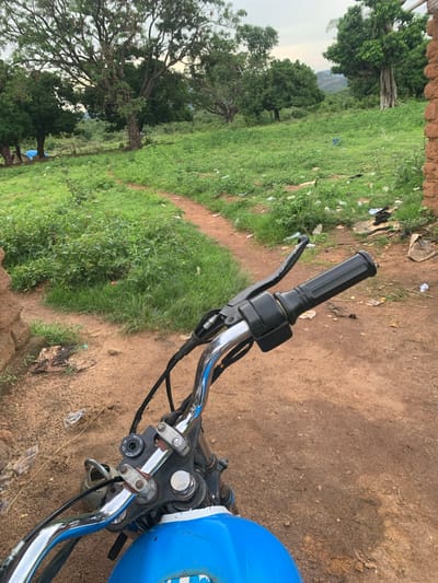 This media file is a first-person perspective shot from a motorcycle, looking forward and slightly to the left. The scene is outdoors in a rural setting, likely in Buga da Gindi, Nigeria, with a path winding through grassy terrain.

The foreground is dominated by the motorcycle's handlebars, which are chrome and black. A black grip is visible on the right side, along with a brake lever and a control unit. The blue fuel tank of the motorcycle is also visible on the right.

The background features a vibrant green, grassy landscape with scattered trees. Some of these trees are large and mature, with thick branches. In the distance, a hint of mountains or hills can be seen under an overcast sky. The ground beside the path is dry, dusty soil, with some scattered litter, including plastic bags and debris.

The time of day appears to be overcast, suggesting it might be morning or late afternoon, or possibly a cloudy day. The overall atmosphere is natural and somewhat unkempt, indicative of a rural or undeveloped area. No people are visible in the image.

The only visible text is on a circular control unit on the handlebar, which has settings for "IGNITION," "LOCK," "ON," and "OFF."