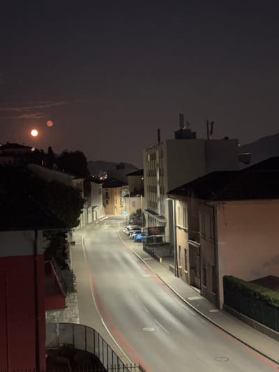 The image depicts a nighttime view of a street in Balerna, Switzerland. A paved road with red markings runs through the center of the frame, curving to the right. Several buildings line the street, varying in height and architectural style. Some buildings have visible windows and balconies. Cars are parked along the side of the road. In the sky, there are two celestial bodies that appear to be the moon, one larger and brighter than the other, tinged with orange. The overall atmosphere is that of a quiet, residential street at night, illuminated by artificial lights from the buildings and street lamps, with a notable presence of the moon. The surrounding landscape suggests a town nestled within a valley, with hills or mountains visible in the distant background.