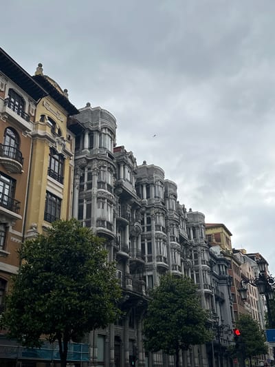 The image captures a view of the historic architecture in Oviedo, Spain, under a cloudy sky. The primary focus is on a row of ornate buildings lining a street. The buildings exhibit a distinct architectural style, with detailed facades featuring rounded bays, balconies with iron railings, and decorative stonework. The building on the left is painted in a warm yellow hue, while its neighbors display grey, lighter stone, and red brick finishes, creating a visually striking contrast. In the foreground, lush green trees with dense foliage are strategically placed, partially obscuring the lower levels of the buildings. A traffic light shows a red signal, and an old-fashioned street lamp with multiple lights stands on the right side of the frame, suggesting an urban setting. The sky is overcast with grey clouds, and a faint silhouette of a bird can be seen flying in the distance. The perspective is from street level, looking upwards towards the buildings, emphasizing their height and architectural grandeur.