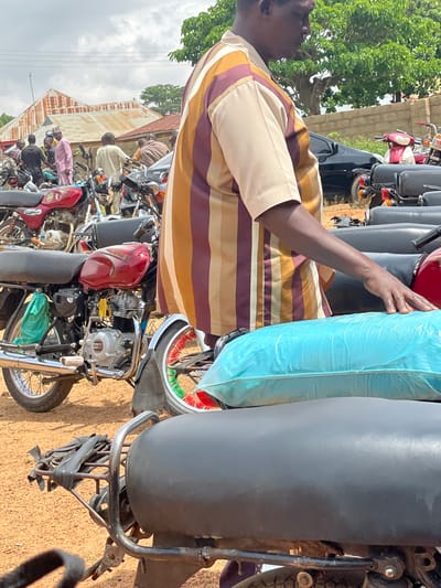 In Kassa, Nigeria, a man in a multi-colored striped shirt stands beside several motorcycles parked on a dirt surface. The man's right arm is extended, resting on a bright blue tarp-covered object. In the background, buildings with corrugated metal roofs are visible, along with trees and a brick wall. Other people are scattered among the motorcycles. A red motorcycle with a green bag attached to it is prominently featured on the left side of the image.