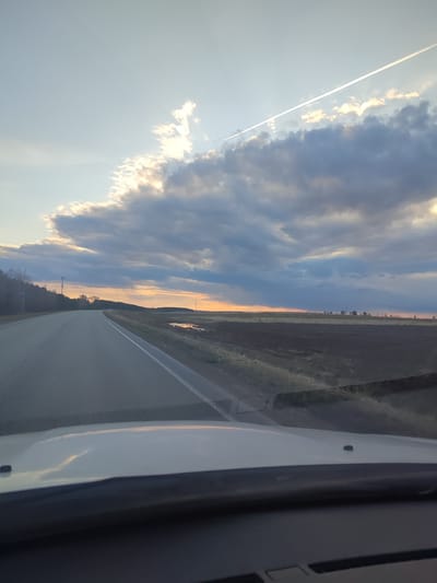 This image captures a serene rural landscape during sunset or late afternoon, viewed from the perspective of a vehicle's front passenger or driver. A two-lane asphalt road with a solid white shoulder line and a broken center line extends into the distance, leading towards a gentle curve. To the left, a dense forest lines the road, while to the right, dark, tilled fields stretch out, featuring several reflective puddles suggesting damp ground.

The sky is a prominent feature, filled with an array of clouds. Some clouds are dark and heavy, while others are illuminated from behind by the low-lying sun, casting warm, golden light across their edges and the distant horizon. A long, straight white contrail diagonally crosses the upper sky. The sun itself is visible as a bright glow near the horizon, partially obscured by clouds.

The bottom of the frame shows the white hood and dark dashboard of the vehicle from which the photograph was taken. The scene evokes a sense of quiet travel through the countryside of Perevoznoye, Russia, as daylight fades. No people or visible text are present in the image.