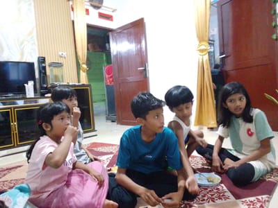 In a room in Gampongjawa, Indonesia, five children are seated cross-legged on a patterned rug. The child on the left wears a pink shirt and skirt, holding a snack. Next to them is a child in a gray shirt. In the center, a child in a blue t-shirt looks to the right. Beside them, another child wears a white tank top. On the right, a child in a teal and white shirt is talking. A blue plate with food sits on the rug. Behind the children, a cabinet supports a TV and speaker. A wooden door is open. Curtains are visible in the background.