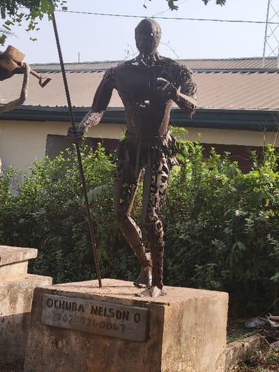 A full-body metal sculpture stands outdoors on a weathered concrete pedestal. The dark, reddish-brown sculpture, constructed from welded metal pieces, depicts a human-like figure in a dynamic standing pose, with its left leg slightly forward. The figure holds a long, thin spear or staff vertically in its left hand, and its right hand, bent at the elbow, holds a small, indistinct object near the chest. A skirt-like garment, also composed of metal fragments, is present around the figure's waist. A rectangular, dark gray plaque is affixed to the front of the concrete pedestal, inscribed with "OCHUBA NELSON O" on the upper line and "02020034-0047" on the lower line. Lush green foliage, primarily bushes, surrounds the base of the pedestal and extends into the background. Behind the vegetation, a building with a light-colored wall, dark trim, and a brown corrugated metal roof is visible. To the left, a portion of another metal sculpture, possibly depicting a figure holding a tool resembling a shovel, is partially visible. The sky above is light blue and clear, with some tree branches overhead on the left. This scene is located in the city of San Martín, Colombia.