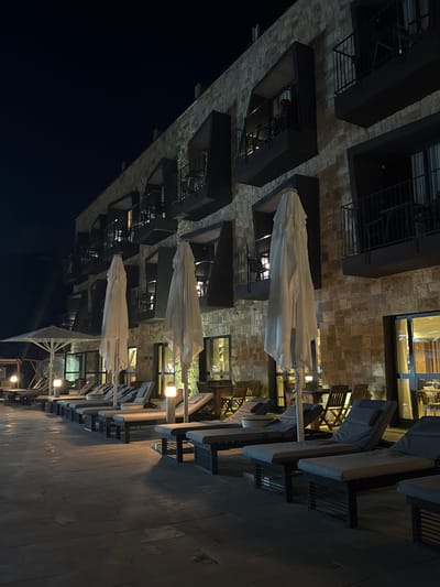 An exterior nighttime shot depicts a modern hotel building with balconies. In the foreground, rows of empty lounge chairs and closed beach umbrellas are set up on a stone patio, suggesting a poolside or outdoor seating area. Lights are visible, illuminating the area. The building's facade is constructed of stone. The scene appears tranquil and uninhabited, typical of a quiet evening at a resort. The location is identified as Tirat Karmel, Israel.