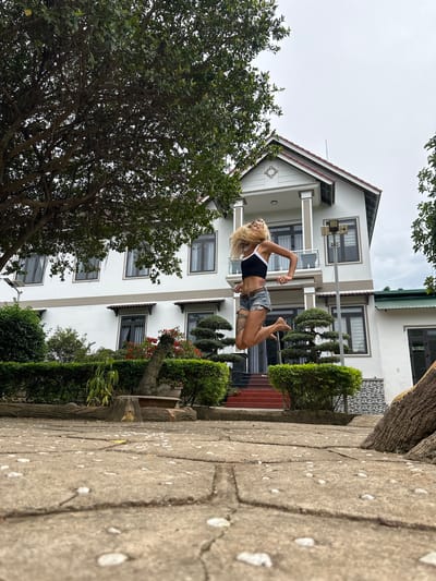 An outdoor, eye-level shot captures a woman mid-air, jumping in front of a multi-story white house with a red-tiled roof. She has blonde hair, wears a black crop top with white trim, and denim shorts. Her body is angled forward, arms extended, with legs bent. The house features several windows with gray frames and curtains, and a covered balcony on the second floor. A large tree with lush green leaves is on the left, and manicured bushes and potted plants are in front of the house. The foreground consists of a paved walkway made of large, square concrete tiles, some of which are uneven. The scene appears to be in a residential area in Thôn 2, Vietnam, judging by the architecture and landscaping. The sky is overcast, suggesting a cloudy day.