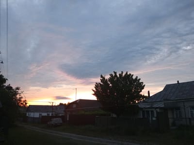 The image depicts a rural village scene at dusk, possibly in Shostka, Ukraine, as indicated by the provided metadata. The sky is filled with a gradient of colors, transitioning from a pale orange and yellow near the horizon to shades of gray and light blue higher up, with wispy clouds present throughout.
In the foreground, a dirt road winds through the scene, bordered by overgrown grass and vegetation on both sides. Several houses with dark, sloped roofs are visible. The house on the right has a visible house number "13" on its facade. Some of these houses are partially obscured by trees and fences. A dark-colored SUV is parked on the left side of the road.
A large, leafy tree stands prominently in the middle ground, silhouetted against the sky, with its branches casting shadows. Power lines are strung across the upper left portion of the image, with a lamppost visible hanging from one of the lines. The overall atmosphere is serene and quiet, characteristic of a rural evening.