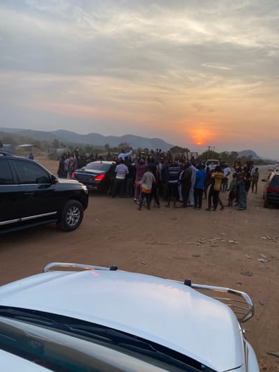 A large crowd gathers on a dirt road as the sun sets. Distant mountains form the horizon under a cloudy sky tinged with orange. Several vehicles are parked along the road, including a black SUV and a white car, partially in the frame. The focus is on the people silhouetted against the bright sunset.