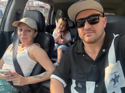 A family of three, a man, a woman, and a young girl, are taking a selfie from inside a car. The man is in the foreground, wearing a black collared shirt, a baseball cap, and sunglasses. He is looking at the camera with a slight smile. To his left, the woman, wearing a beige baseball cap and a white tank top, is also looking at the camera, holding a smartphone in her hands. Behind them, the young girl with long brown hair is seated and looking towards the camera with a sweet expression, resting her chin on her hand. The interior of the car is visible, with black leather seats and seatbelts. Through the car windows, a building with multiple floors and windows can be seen, suggesting an urban or suburban setting. The lighting suggests it is daytime, possibly late afternoon due to the warm, soft light. The overall mood is casual and familial. There is visible text on the man's shirt: "LOS ANGELES", a star graphic, "ESTD", and "CALIFORNIA". There is also text on a tag on his shirt: "AOMD YUEMLD FASHION".