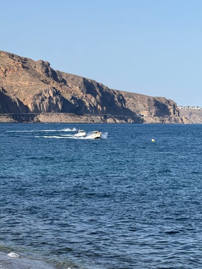 This media file depicts a scenic coastal view with a dramatic mountain backdrop and a vibrant blue sea. On the left side of the frame, a rugged, rocky mountain rises steeply, its surface textured with shadows and highlights suggesting the late afternoon sun. A narrow, winding road or path is visible ascending the mountainside, with a few sparse buildings perched near the summit, hinting at a settlement or resort.
The right side of the image is dominated by the deep blue of the sea, its surface agitated with numerous small waves and ripples, indicating a breezy day. A small, yellow object, possibly a buoy or a piece of debris, floats on the water's surface. Further out, a white motorboat is in motion, leaving a prominent white wake that trails behind it. A single person is visible on the boat, and another person appears to be riding a jet ski behind the boat, creating a spray of water. The overall scene suggests a leisurely day at the coast, with recreational activities taking place on the water.
The lighting suggests it is late afternoon, with the sun casting long shadows on the mountain. The clear sky indicates fair weather. The location cues, such as the type of mountain and the coastal setting, combined with the provided context, point to Aguadulce, Spain. There is no visible text in the image. The image evokes a sense of adventure and relaxation.