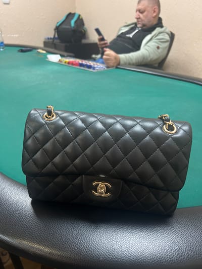 The image shows a black quilted Chanel handbag with gold hardware resting on a black leather surface. In the background, a person is seated at a green poker table, holding a dark object. There are poker chips visible on the table. The setting appears to be indoors. There is no information available that indicates it is in the city of Smederevska Palanka Municipality, Serbia.