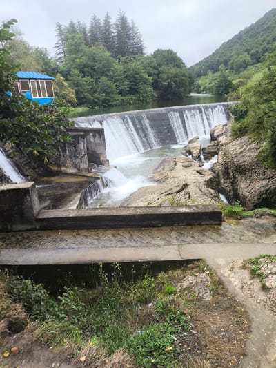 A wide shot captures a cascading waterfall in Gachedili, Georgia, with lush green trees and a mountainous backdrop under an overcast sky. A blue structure with large windows sits on the left side of the dam. The water flows over a concrete dam, creating a frothy white spray that tumbles down into the rocks and the river below. The surrounding landscape is densely forested, with the mountains rising steeply in the distance. The image appears to be taken from a slightly elevated position, looking down towards the waterfall and the river.