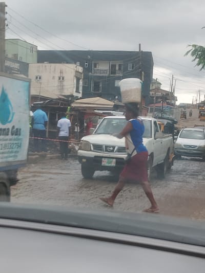 A woman is seen walking across a wet, muddy road in what appears to be a busy street market in Iju, Nigeria. She is carrying a large white bucket filled with what looks like grain or other foodstuff on her head. She is wearing a blue t-shirt and a maroon skirt, with sandals on her feet.
In the background, there are several vehicles, including a white pickup truck, a white sedan, and various other cars and motorcycles, suggesting a trafficked area. Buildings line the street, some appearing to be shops or stalls with makeshift roofs. The ground is visibly wet and appears to be covered in puddles, indicating recent rain or ongoing damp conditions.
The sky is overcast, suggesting a cloudy or possibly rainy day. The scene has the feel of a typical market day, with people engaged in various activities, though they are not clearly discernible due to the image's resolution. There is a partial view of a truck on the left side of the image with some text on it. Visible text on the truck includes "anna Gas" and a phone number "149332754". The overall atmosphere seems to be one of everyday commerce and movement.