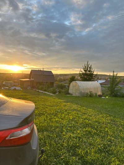 The image depicts a rural or suburban landscape at what appears to be sunset or sunrise in Bol'shaya Mos', Russia. In the immediate foreground, the rear left section of a dark grey Kia Rio sedan, identifiable by the "RIO" emblem, is visible, with its taillight and trunk reflecting strong golden sunlight. A tow hitch is attached to the vehicle's underside. The foreground and midground consist of a vibrant green, sunlit grassy field containing small white flowers. In the midground, a two-story, rustic wooden house with a dark metal roof stands, featuring a wooden ladder propped against its side and a small attached porch. To the right of the house, a clear, tunnel-shaped greenhouse made of transparent material is situated, flanked by a tall, slender pine tree and other sparse vegetation. The background reveals a residential area with numerous houses and structures extending towards a distant horizon, with faint utility lines visible. The sky transitions from a bright, golden glow on the left horizon, where the sun is prominent, to a predominantly cloudy expanse of grey and light blue hues overhead. No people or specific actions are discernible.