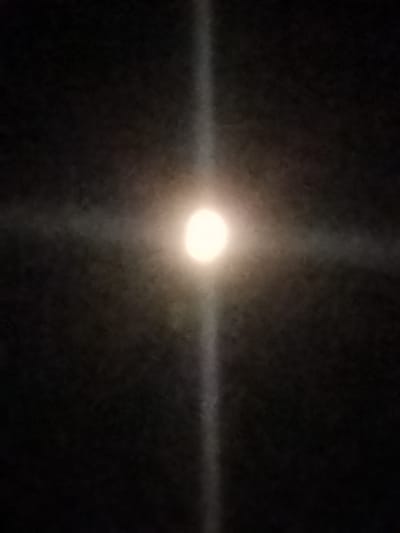 The image is a dark, grainy photograph taken at night. It shows a bright, circular object in the center, which appears to be the moon or a strong light source. A faint cross of light extends outwards from this central object, suggesting a lens flare effect or light diffracted by atmospheric conditions. The background is black, indicating a night sky or a dark interior space with no other discernible objects or features. There are no people, animals, or specific landmarks visible in the image, making it impossible to determine a location within Kostiantynivka, Ukraine, or any other geographical context.