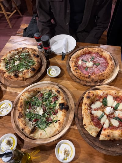 This image displays an indoor dining scene at a restaurant in Ban Tin That, Thailand. A light-colored wooden plank table serves as the primary surface for four individually prepared pizzas, each presented on a unique round wooden serving board.
From the top-left, proceeding clockwise:
1. A round pizza features a robust, slightly charred crust, topped with dark caramelized ingredients, fresh arugula, and several white dollops, possibly of cheese or a creamy sauce.
2. A round pizza with a similarly charred crust is adorned with thinly sliced red cured meat (e.g., salami), small green basil leaves, and white dollops.
3. A roughly square-shaped pizza with a prominent, slightly irregular crust is covered in red tomato sauce, substantial pieces of fresh mozzarella cheese, and green basil leaves. One slice is partially separated from the main pie.
4. A roughly oval-shaped pizza, also with a dark-edged crust, is topped with cured meat (e.g., prosciutto), fresh arugula, and white dollops.
Accompanying the pizzas are several ancillary items:
* Two salt and pepper grinders are positioned between the top two pizzas, one labeled "McCormick Black Peppercorn."
* An empty white ceramic plate containing a folded white napkin, a silver fork, and a knife is situated near the pepper grinder.
* Four small white ceramic bowls, three visible in the foreground and one on the left, each contain a white substance (likely labneh or a soft cheese) drizzled with olive oil and sprinkled with a dry spice.
* A clear glass bottle of olive oil with a pourer top is visible in the bottom-left corner.
In the upper background, partially visible, a person is seated at the table, wearing a dark brown long-sleeved shirt or jacket, indicating their presence as a diner. The overall lighting is warm, suggesting an evening or intimate atmosphere.