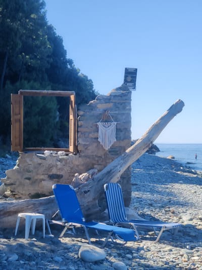 This image captures a beach scene in the Municipal Unit of Evrymenes, Greece. The focal point is the partially ruined stone structure of a building with an open wooden window frame. A large piece of driftwood leans against the structure. In front of the ruin, two lounge chairs, one solid blue and the other blue and white striped, are set up on a pebble beach. A small white plastic stool sits to the left of the chairs. Behind the chairs and driftwood, the blue sea stretches to the horizon, with a single person visible wading in the water. Lush green trees line the backdrop behind the ruin. A macrame decoration hangs on the stone wall, and a light fixture is mounted on top. The sky is clear and blue.