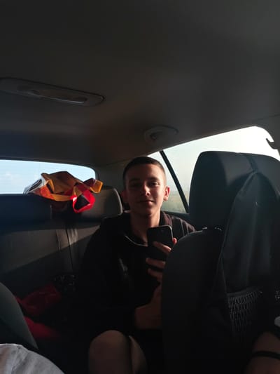 A young male is seated in the back seat of a car. He is holding a smartphone. The car's interior is visible, including the seats, ceiling light, and a shopping bag behind him. The setting appears to be Bogdanovo, Bulgaria, indicated by the view through the car window showing landscape with plants and sky.