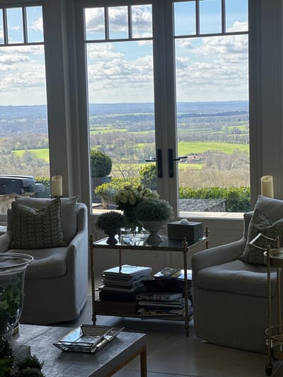 This image depicts an interior living space in Sevenoaks, United Kingdom, overlooking a panoramic rural landscape. The scene is presented from a low-angle perspective, showing a room furnished with two light-colored upholstered armchairs, each adorned with a green patterned cushion.
A central coffee table is partially visible in the foreground, displaying a geometric glass and metal tray and a large clear glass vase containing green foliage. To the left, a cream-colored pillar candle rests on a ledge near the window. To the right of the scene, another cream-colored pillar candle is visible on a similar surface.
Between the two armchairs, a two-tiered brass-framed side table with a glass top holds a vase of white and green floral arrangements, a smaller potted plant, and a rectangular dark-colored tissue box. Below this, on the lower shelf, a stack of magazines and books is visible. Another brass-framed serving cart is partially visible to the right, adjacent to the right armchair. The floor is covered in light-colored tiles.
The prominent feature is a series of tall, multi-paned windows and a central glass door section with dark handles, offering an expansive view of the exterior. The landscape beyond consists of rolling green hills, fields, and scattered deciduous trees with bare branches, extending towards a distant horizon line. A small cluster of buildings, possibly a farm or village, is discernible in the middle distance. The sky is bright blue with scattered white cumulus clouds. A very faint, small airplane is visible high in the sky through the windows. Directly outside the windows, a gabion wall is visible, along with potted plants and what appears to be outdoor furniture or a grill on a stone patio.
No people are present in the image. The lighting is natural, originating from the large windows, casting parts of the interior into shadow.