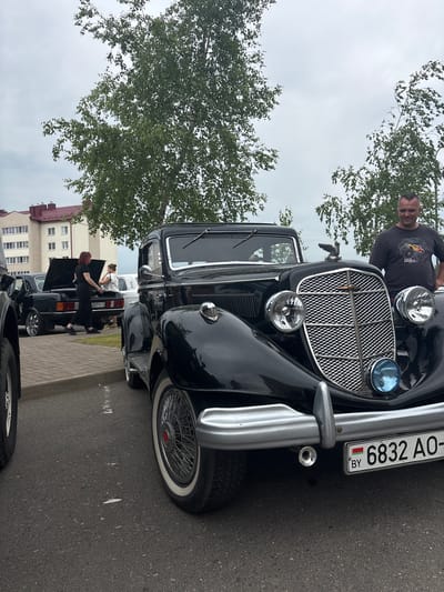 In Uzda, Belarus, a man in a dark t-shirt with a graphic print stands next to a vintage black car. The car, with its chrome grille, round headlights, and a blue spotlight, is the primary subject. The man is looking down and slightly to his right. Behind the car, a group of people are gathered around another black car with an open trunk, and further in the background, a multi-story building with a red roof is visible. Trees with green foliage frame the scene. The ground is paved with asphalt and cobblestones. The sky is overcast.