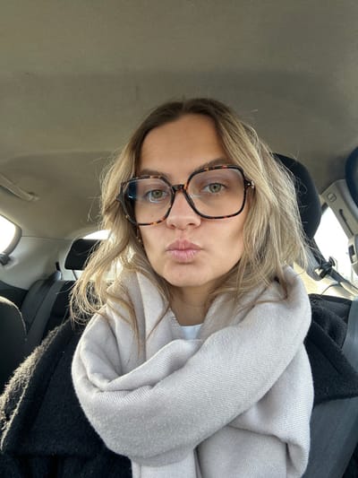 The image displays a female individual seated within the interior of a vehicle in Veauche, France. The subject is depicted in a close-up view, facing the camera directly with a deliberate pouting expression. She has light brown, wavy hair and wears large, tortoise-shell patterned glasses, through which her green eyes are visible. A light beige, possibly cashmere or wool, scarf is wrapped around her neck and shoulders, partially obscuring a dark-colored garment worn underneath. The vehicle's interior is visible in the background, featuring a light-colored headliner and dark upholstered rear seats. Bright natural sunlight illuminates the scene from the right side of the frame, indicating a daytime setting.
