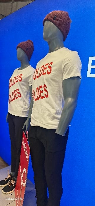Two mannequins stand against a blue background. Each wears a maroon beanie, white t-shirt with the word "SOLDES" printed in red, black pants, and black shoes. A red sign with a white circle is positioned near the bottom mannequin, partially obscuring the shoes. The background is a bright, solid blue.