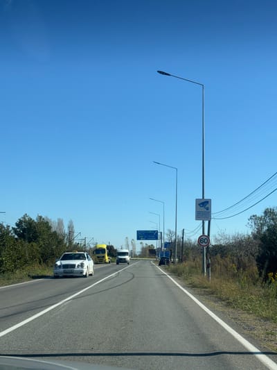 A street view shows a road with several vehicles driving on it. A white car is in the foreground. Road signs and light poles are on the side of the road. The sky is clear blue. Trees and vegetation line the road. The road surface is gray and shows tire marks.