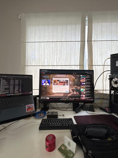Here is a description of the image:

The image shows a desk setup in front of a window with horizontal blinds. A desktop computer tower is visible at the top, along with a monitor displaying a YouTube page. Below the monitor is a laptop with its screen displaying file folders. To the right of the monitor are a mechanical keyboard, a small plastic figurine, a red cylindrical object labeled "Rolling Circus", a green and white box labeled "Stamps World", a black backpack, and a dark mousepad. The environment is a room with a neutral-colored wall behind the window.