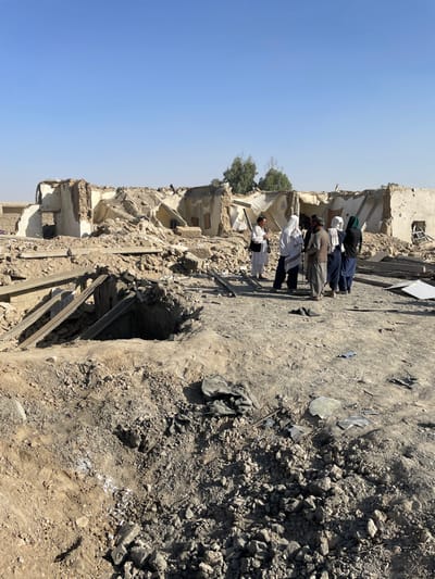 The image displays a scene of extensive structural destruction in Spin Boldak, Afghanistan, under a clear, light blue sky. In the immediate foreground, a significant, irregularly shaped crater, approximately 3 meters in diameter, is visible in the earth, revealing exposed structural elements resembling rebar and wooden beams within its excavated cavity. The ground throughout the scene is uneven, covered with a dense layer of pulverized earth, rubble, concrete fragments, and scattered debris. A crumpled dark fabric object rests on the ground to the right of the crater.
In the mid-ground, a group of six individuals is positioned amidst the debris. From left to right: one individual in white traditional attire, wearing sunglasses and carrying a dark shoulder bag, faces the group. Adjacent, another individual in a white head covering holds a dark, elongated object, possibly a microphone or recording device, oriented towards the cluster. Next is a male individual dressed in a light-colored traditional outfit and a dark waistcoat. The remaining three individuals are attired in traditional garments with head coverings, two white and one dark green, clustered in close proximity. Their stance and interaction suggest conversation or observation of the site.
The background is dominated by the remnants of severely damaged buildings. These structures, appearing to be single-story or multi-story constructions, have suffered significant collapse, with large sections of their roofs and upper walls missing. The remaining wall segments, constructed of light-colored material, are fragmented, displaying numerous holes and exposed internal structural elements. Piles of rubble form the base of these demolished structures. Distant, sparse green trees are visible on the horizon behind the destruction. The overall environment suggests a dry, arid climate.