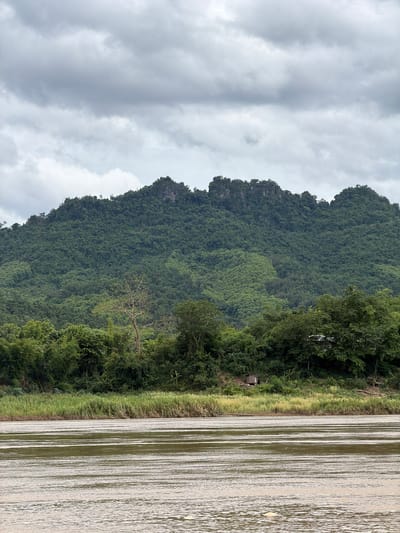 A wide, muddy river flows across the foreground of the image. In the middle ground, a grassy bank is lined with lush green trees and vegetation, with a small wooden hut visible amongst the foliage. Towering in the background is a steep, heavily forested mountain with rocky outcrops at its peak. The sky above is overcast with thick, grey clouds. The scene appears to be a natural landscape, likely in a rural or undeveloped area.