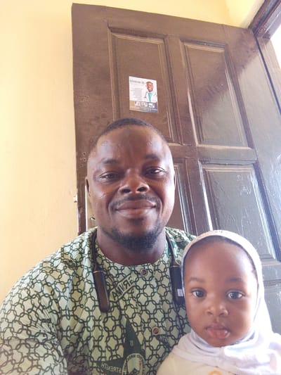 A selfie taken by a dark-skinned man with a close-cropped haircut and a thin beard. He is wearing a patterned green and white shirt and a stethoscope. He is holding a young child with a white head covering and scarf. Behind them is a brown paneled door with a business card attached to it featuring a man in a white coat with a stethoscope. The background includes a yellow wall. This image was taken in the city of Dungu, Ghana.