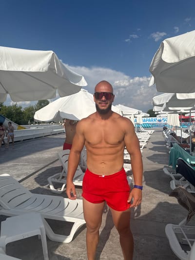 A man, appearing to be in his late 20s to early 30s, stands facing the camera in an outdoor setting. He is shirtless, wearing red swim trunks, sunglasses, and a blue wristband. His muscular physique is visible. The background suggests a water park or similar recreational area, with rows of white lounge chairs and large white umbrellas. In the distance, water slides and other park structures are visible, along with a blue sky with scattered clouds. The man is the primary subject, positioned centrally in the frame. The overall scene is bright and appears to be during daylight hours. The location is identified as Ivanyane, Bulgaria.
