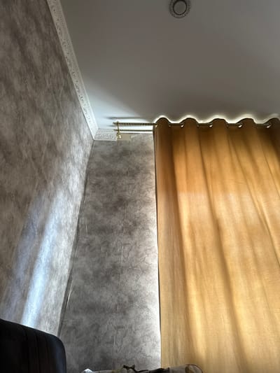 The image depicts an interior scene, likely a room, focusing on a corner where two walls meet a white ceiling. The walls have a textured, mottled gray finish. A decorative white molding runs along the junction of the walls and ceiling. A curtain rod with a golden finish is installed below the molding, supporting a sheer, golden-brown curtain. The curtain is drawn, partially obscuring the view of the wall behind it, and allows light to filter through, creating a soft glow. A recessed circular light fixture is visible in the ceiling. The lower left portion of the image shows a dark upholstered object, possibly furniture. The scene appears to be a residential interior in Nazran, Russia.