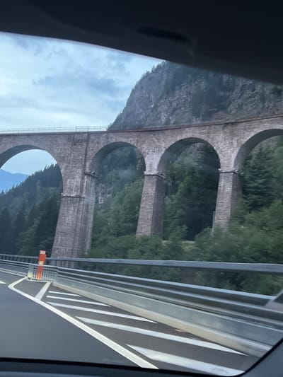 The image captures a stone viaduct with multiple arches, likely a railway bridge, spanning across a lush, green valley. The viaduct is constructed from large, rough-hewn stone blocks and features robust, tapering pillars supporting the arches. The top of the viaduct appears to have a railing, and what looks like railway tracks can be seen. The surrounding environment is densely forested with tall, dark green trees. A steep, rocky mountain slope rises behind the viaduct, with a cloudy sky overhead. In the foreground, a modern road with white lane markings and a metal guardrail is visible, suggesting the photograph was taken from a vehicle on a road below or adjacent to the viaduct. An orange traffic marker is present on the side of the road. The location is identified as a viaduct in Les Houches, France.