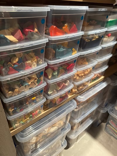 Rows of clear, plastic containers fill a wooden drawer. Each container is filled with various colorful objects, possibly fragments of toys or art supplies. The containers are neatly aligned, creating a textured, organized view. Some containers have lids. The lighting highlights the textures of the drawer and the contents of the containers.