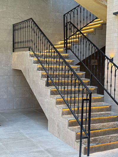 The image shows an exterior staircase with a modern design, located in the city of Ayn al Basha, Jordan. The staircase features grey stone-tiled steps and risers, each illuminated by a warm, yellow LED strip light. A black wrought-iron railing with an ornate, arched design runs along both sides of the stairs. The walls surrounding the staircase are clad in large, grey stone tiles, creating a contemporary and sophisticated aesthetic. The floor at the base of the stairs is also tiled with grey square tiles. The lighting suggests it is either dusk or night. There are no people visible in the image.