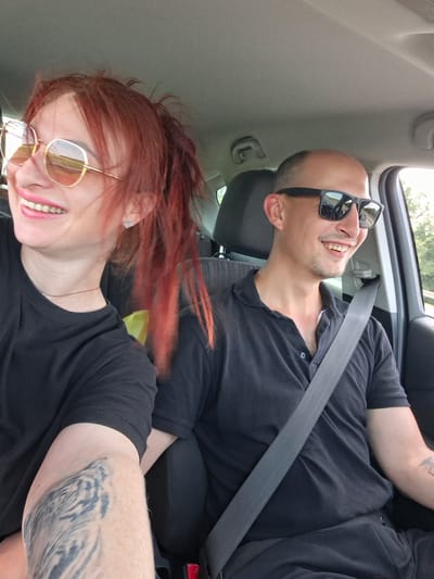 In the image, two individuals, a woman with red hair and sunglasses and a man with sunglasses, are seated in the front seats of a car, seemingly in motion. The woman is on the left, facing forward and smiling, with her hair pulled back in a messy ponytail. She is wearing a black t-shirt and gold-rimmed sunglasses with orange lenses. The man is on the right, also facing forward and smiling. He is wearing a black polo shirt and black sunglasses. A gray seatbelt is visible across his chest. The interior of the car, including the dashboard and seats, is visible. The background outside the car suggests a rural or suburban setting with trees and a road. The overall impression is a snapshot of a couple enjoying a drive, possibly in or near the city of Navadvyorskoye, Belarus, given the context.