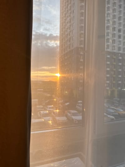 The image depicts a view from a window in Drozhzhino, Russia, at sunset. The foreground is dominated by a brown curtain on the left, partially obscuring the view. Through the window, two tall residential buildings are visible, one on the right and another further back. The sky displays a warm sunset with clouds. The sun, a bright orb with a lens flare, is setting in the distance. Below the buildings, a parking lot is filled with cars. The scene is bathed in the golden light of the setting sun, which also reflects off the window, creating a hazy and somewhat abstract effect. The window itself appears to have some dust or grime, further contributing to the diffused lighting.