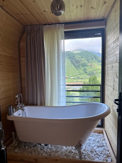 The image shows a clawfoot bathtub in a wooden room with a large window overlooking a mountainous landscape. The bathtub is white with silver fixtures and stands on a bed of white stones. A beige curtain is visible to the left of the window. The window reveals a green mountain range under a cloudy sky. This scene appears to be located in Sioni, Georgia, a village known for its natural beauty and mountainous surroundings. There are no people or animals visible in the image.