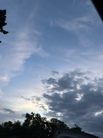 The image shows a wide-angle view of a cloudy sky with trees in the foreground. The sky is a mix of light blue and dark gray clouds, suggesting either an approaching storm or the aftermath of one. Wispy cirrus clouds are visible against the lighter blue portions of the sky, while thicker, more ominous cumulonimbus clouds dominate the lower half of the frame.
In the lower part of the image, the dark silhouettes of trees are visible against the sky. These appear to be deciduous trees, with dense foliage. To the right, a portion of a light-colored, possibly metallic, object is visible, curving upwards. This object appears to be man-made, and its shape suggests it could be part of a vehicle or some outdoor equipment.
There are no people or animals visible in the image. The scene is static and appears to be taken from a ground-level perspective, possibly from a backyard or a park. The overall impression is one of a natural landscape under a dramatic sky.
Without additional information, it is impossible to pinpoint the exact location within Magdeburg, Germany. However, the presence of trees and the general sky conditions are consistent with a setting in a suburban or semi-rural area of the city, or perhaps a park within Magdeburg. The man-made object in the foreground adds an element of modern life to the otherwise natural scene.