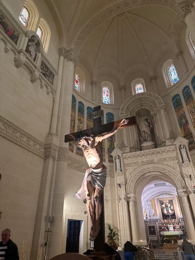 This image depicts the ornate interior of a church located in Għarb, Malta. A large, realistic sculpture of Jesus Christ on a dark wooden crucifix is positioned centrally in the foreground, angled slightly right. The sculpture shows visible wounds on the hands, feet, and torso with simulated blood, a crown of thorns, and a white loincloth. The letters "INRI" are visible at the top of the cross.

The church features light-colored stone or plaster walls and high, vaulted ceilings adorned with intricate relief carvings, including geometric patterns. Multiple arched windows with colorful stained glass are visible in the upper sections, admitting light. Below these windows, various religious murals and mosaic panels depicting figures like saints and angels are integrated into the wall design. Ornate, fluted columns with detailed capitals support the architectural structure.

In the background, an elaborately carved archway bears the inscription "VETVS MARIA AEDICVLA." Above this arch, a large white statue of a robed figure stands within a central niche, flanked by smaller white angelic statues. Beyond the archway, a brightly lit altar is visible. The altar features a prominent, gold-framed painting depicting the Virgin Mary surrounded by angels. Numerous lit candles are arranged on tiered stands on and around the altar. Additional white statues are present near the altar.

On the lower left, an adult male subject wearing glasses and a striped shirt is partially visible, facing away from the camera's direct line of sight. On the lower right, two individuals are observed from behind, kneeling or bending forward near candle stands displaying multiple lit candles. A partially open dark wooden door is visible on the right wall. A small wooden cross and some decorative branches are positioned on the lower left wall. The lighting suggests a combination of natural light from stained glass and artificial spotlights and hanging lamps illuminating the interior.