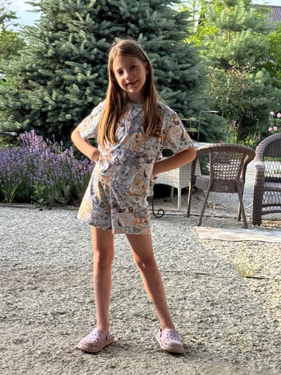 A young girl with long, blonde hair stands in a yard, facing the camera with a smile. She is wearing a light-colored, short-sleeved dress with a pattern of cartoon dogs. The girl is barefoot, with her right foot visible in the foreground. Her left leg is extended backward.

The setting appears to be an outdoor patio or garden area. In the background, there are large green evergreen trees and a patch of purple lavender flowers. Behind the girl, a wicker chair and a white fabric are visible, suggesting a relaxed, possibly domestic environment. The ground is covered in small, light-colored gravel.

The lighting suggests it is daytime, likely sunny, casting shadows. The overall mood seems cheerful and casual. The image is oriented vertically, with the girl positioned in the center.