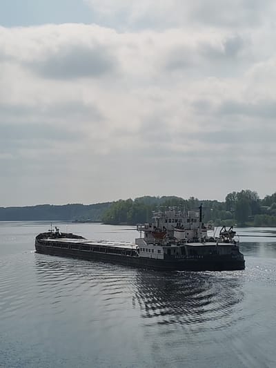 A barge is sailing on a body of water near Glebovo, Russia. The barge, identified as "VOLGA-DON-149," is dark-colored and elongated. It is traveling on a calm body of water, possibly a river or lake. Trees line the distant shoreline, and the sky overhead is overcast with scattered clouds.