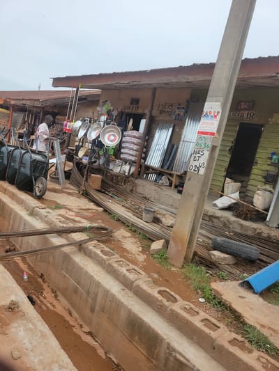 The image depicts a scene in Tangu, Nigeria, likely a marketplace or a construction materials yard. The main subjects are various objects, including building materials like rebar and corrugated metal sheets, as well as household items such as pots and pans displayed for sale. There are also two wheelbarrows, one of which is partially visible, loaded with items. A concrete pillar runs vertically through the left side of the image, supporting an overhead structure that appears to be a shop or stall.

The setting is outdoors, with a reddish-brown dirt ground and evidence of recent rain, indicated by wet patches and mud. The sky is overcast, suggesting a cloudy day. The structures in the background are simple, with corrugated metal roofs and walls. The overall impression is one of a busy, informal commercial area focused on construction and household goods.

Visible text includes signage such as "BLOCK A 17" and "BLOCK A 18," indicating an organized layout of stalls or units. There is also a poster or flyer with the text "PENTECOST FIRE CONFERENCE," featuring a group of people. Other legible text includes brand names like "BUA CEMENT" on bags, and a series of numbers and letters, possibly for identification or pricing: "33 0536 NES BT 029 43".

No specific activity or event is clearly taking place, but the arrangement of goods suggests ongoing commercial transactions. The lighting is diffused due to the overcast sky. The location cues point to Nigeria. There are no prominent emotional expressions to observe, as the focus is on the objects and the environment.