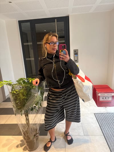 An adult female is depicted taking a mirror selfie in a well-lit indoor hallway or entryway in Nogent-sur-Marne, France. The subject, with blonde hair and black-framed glasses, is wearing a dark grey zippered jacket, a dark undergarment visible at her midriff, black and grey striped knee-length shorts, and black toe-separated flat shoes. White wired earbuds are connected to her magenta-cased smartphone, which she holds with her right hand to capture the image. Her left hand rests on a tall, leafy green plant in a pot, wrapped in clear plastic. A white shopping bag with red markings hangs from her right arm. To her right, a red wall-mounted box with the text "RESERVE POMPIERS" (Firefighters' Reserve) and "APPUYER ICI" (Press Here) is visible, along with a grey intercom panel. The floor consists of large black and white checkered tiles transitioning to lighter grey tiles, with a dark grey ribbed doormat on the far right. The walls are plain white, and the ceiling features white perforated acoustic panels. In the background, a dark grey double door with glass panels reflects ambient light.