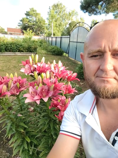 A man with a shaved head and beard takes a selfie in a garden. He is wearing a white polo shirt with red and blue stripes on the collar and sleeves. In the foreground, there are several pink lilies with green stems and leaves. Behind the man, a silver gate and fence are visible, with trees and a building in the background. The scene appears to be a private backyard in a city setting.